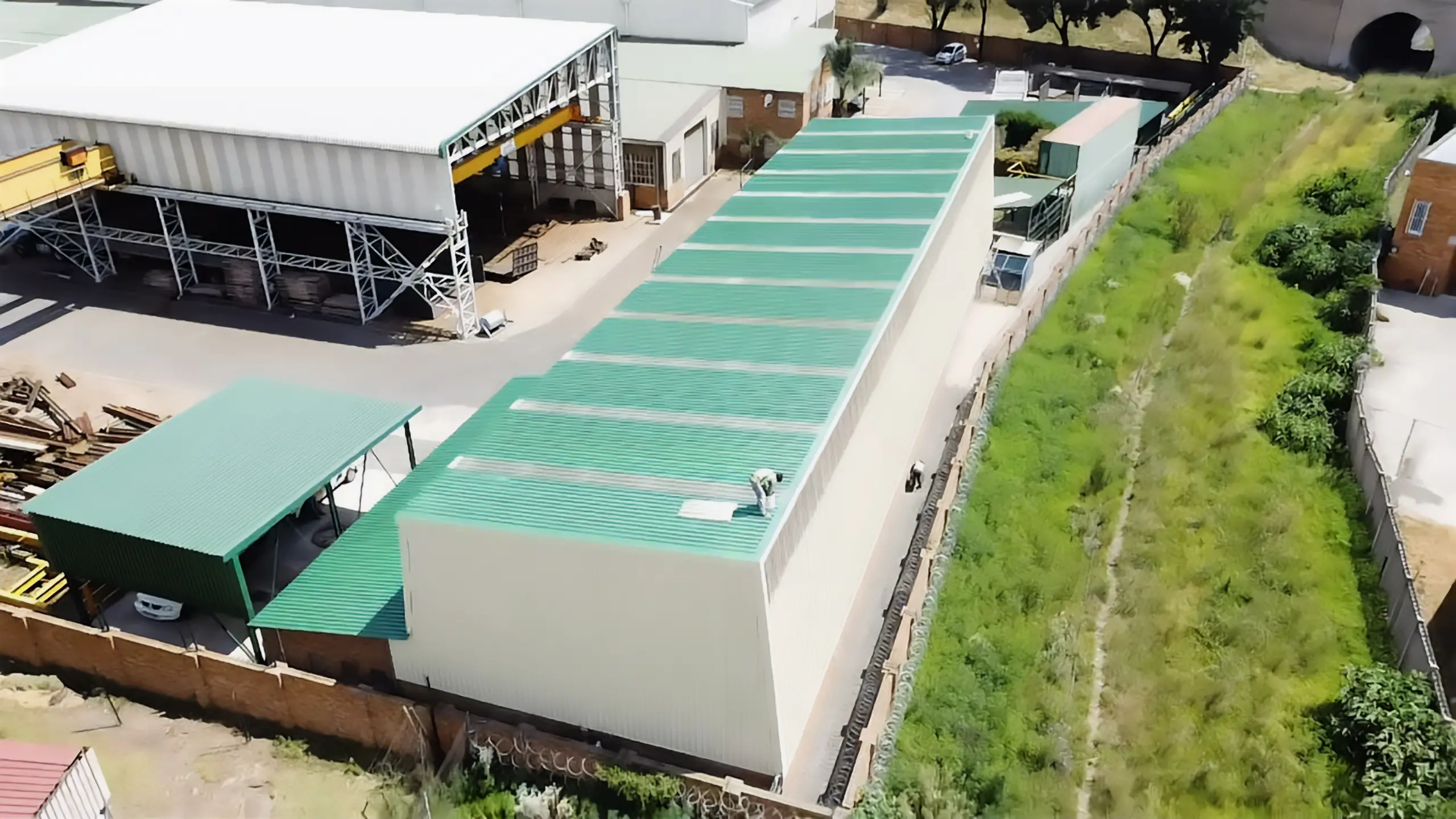 Aerial view of a long industrial building with a green corrugated roof. Workers are seen performing roof refurbishments and maintenance tasks.
