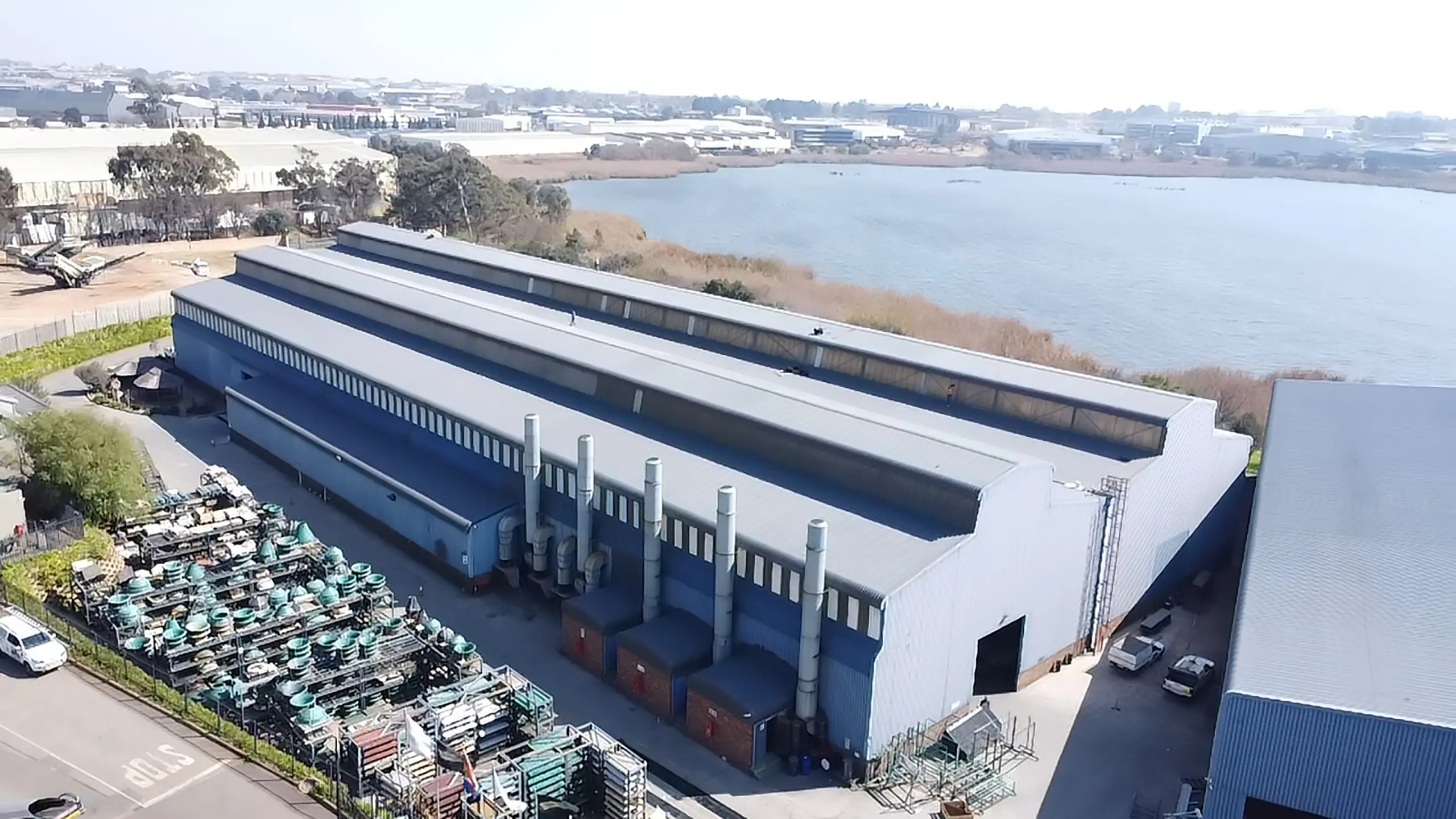 Aerial view of a large blue industrial factory with a long grey roof, next to a lake, suitable for major roof refurbishments.